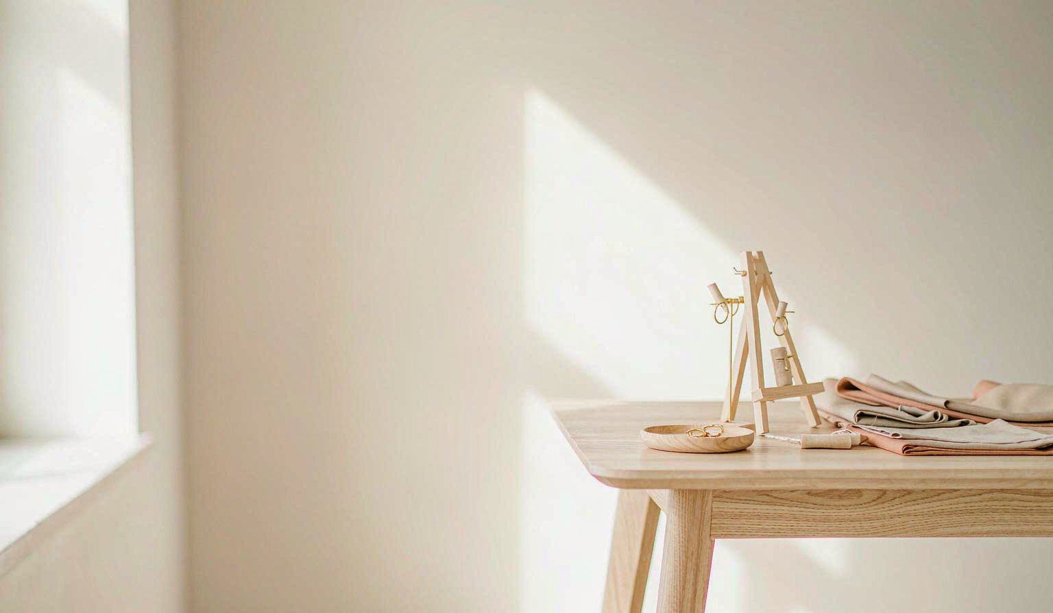 A pale studio interior in daylight: an oak table on the right holds a small jewellery display with rings and folded fabric swatches, against a wall with a soft rectangle of window light